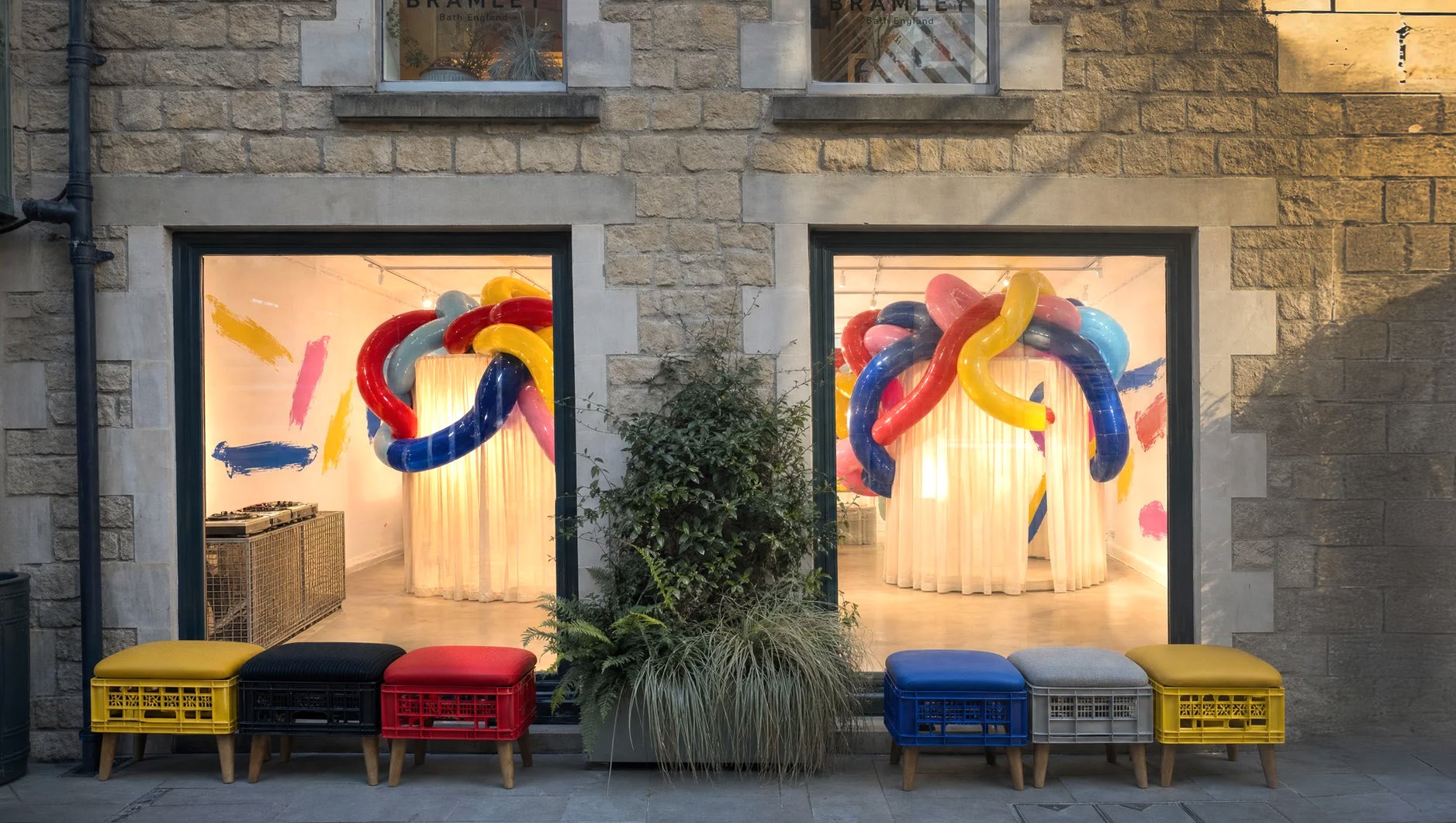 Visualisation of the Shires Yard shopfront at dusk: through both ground-floor windows, a glowing curtained drum wrapped in giant glossy balloon forms, painted marks on the walls and decks on the left; colourful crate stools line the pavement outside