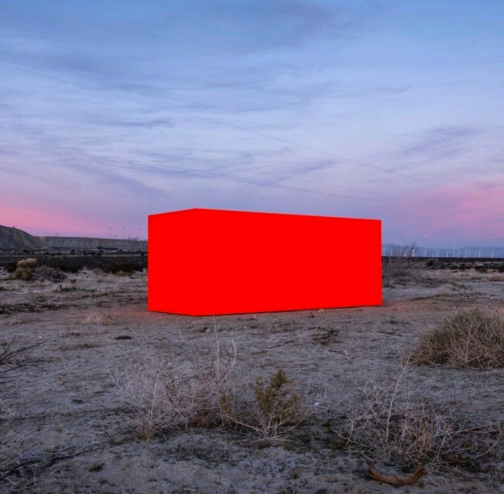 A bright red rectangular block standing alone in a desert at dusk